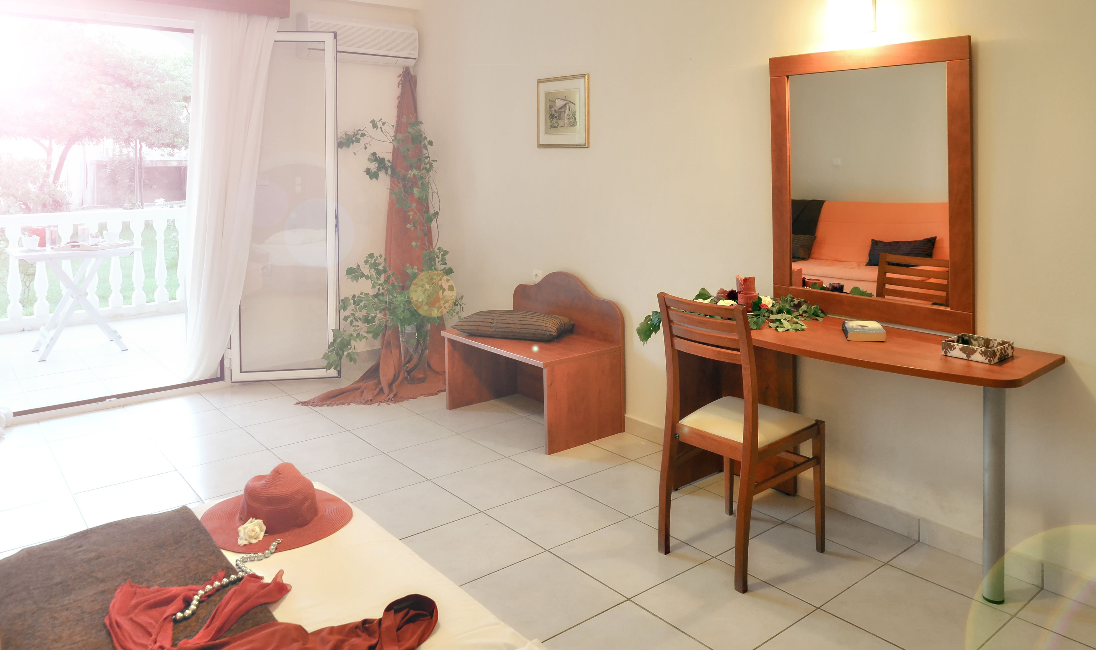 A room with a wooden desk, a chair, a bench, a large mirror, and a view of a balcony with a white railing.