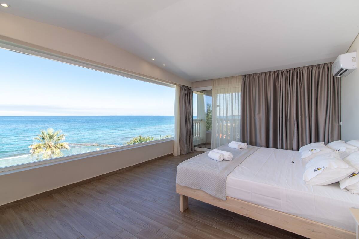 Une chambre avec une grande fenêtre donnant sur la mer, un lit avec du linge de maison blanc et un plancher en bois.