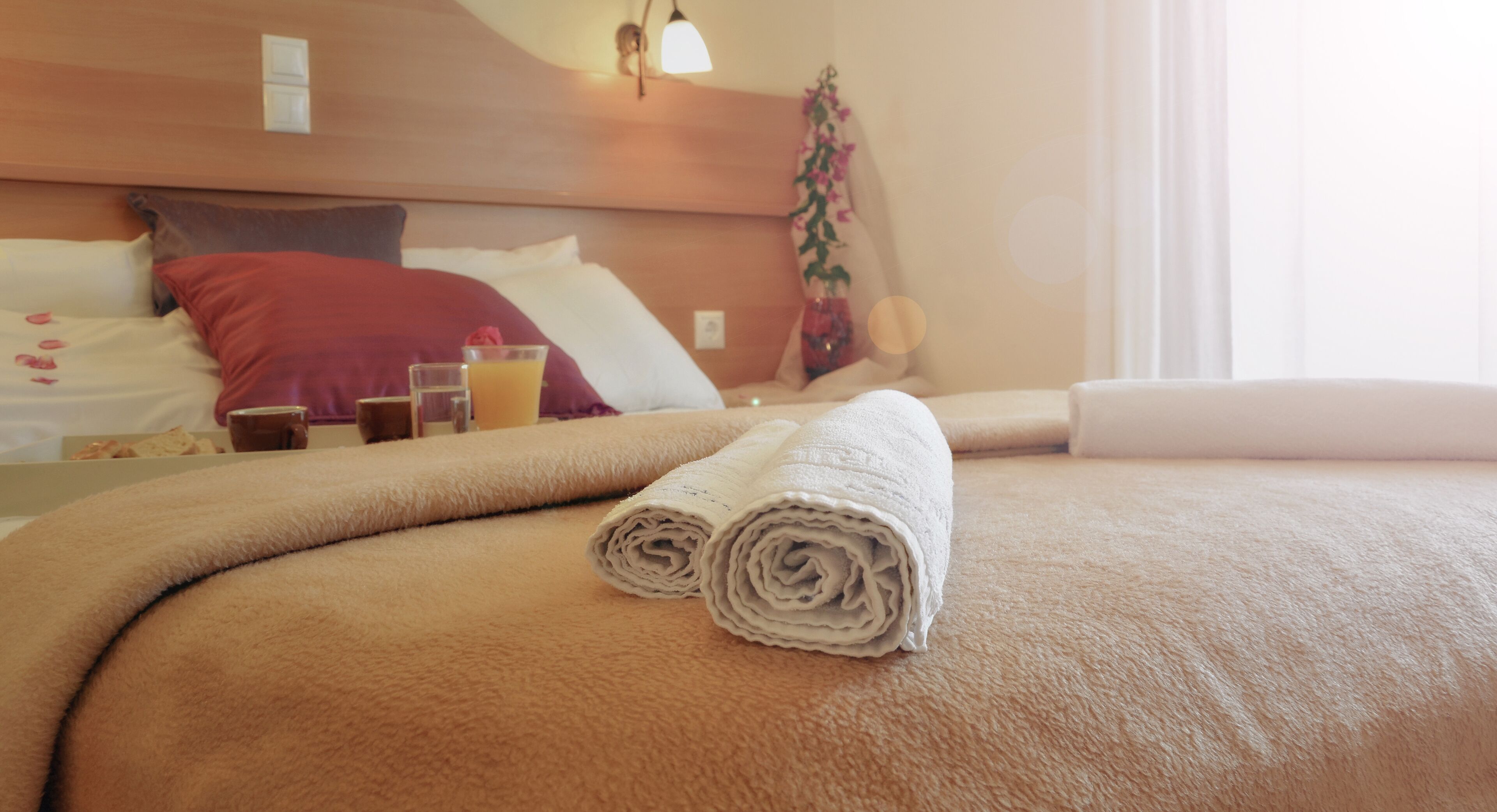 A neatly made bed with rolled towels, a bedside table with a glass of orange juice, and a potted plant.