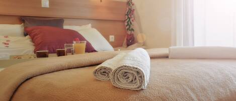 A neatly made bed with rolled towels, a bedside table with a glass of orange juice, and a potted plant.