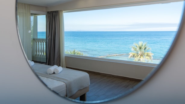 Une chambre avec une grande fenêtre donnant sur la mer, un lit avec du linge de maison blanc et une vue sur les palmiers.
