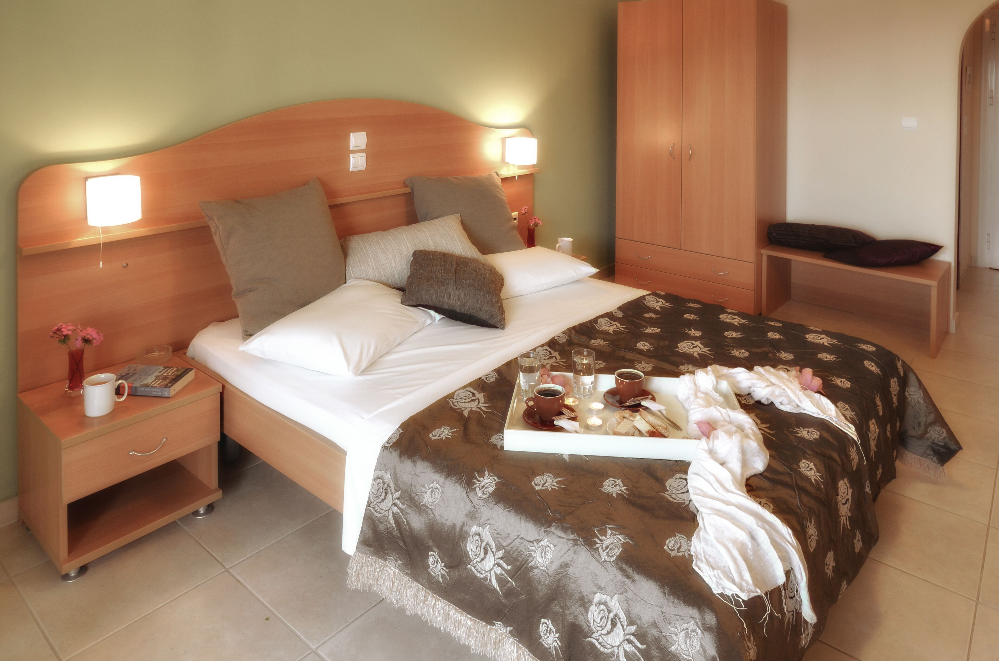 A neatly made bed with a floral bedspread, bedside lamps, a nightstand with a book and a mug, and a wooden wardrobe.
