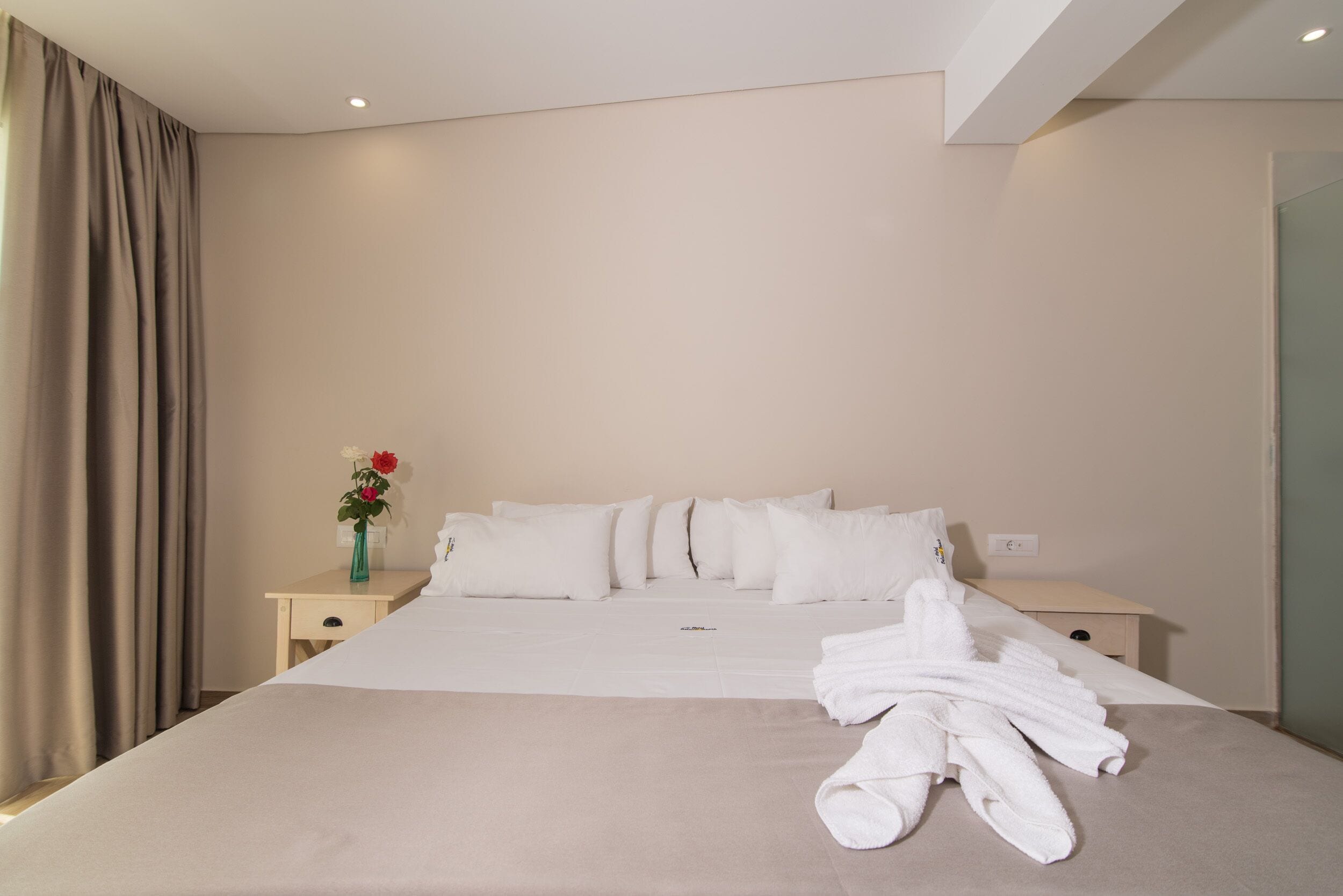 Quarto de hotel com uma cama grande, duas mesas de cabeceira, um vaso com flores e um conjunto de toalhas na cama.