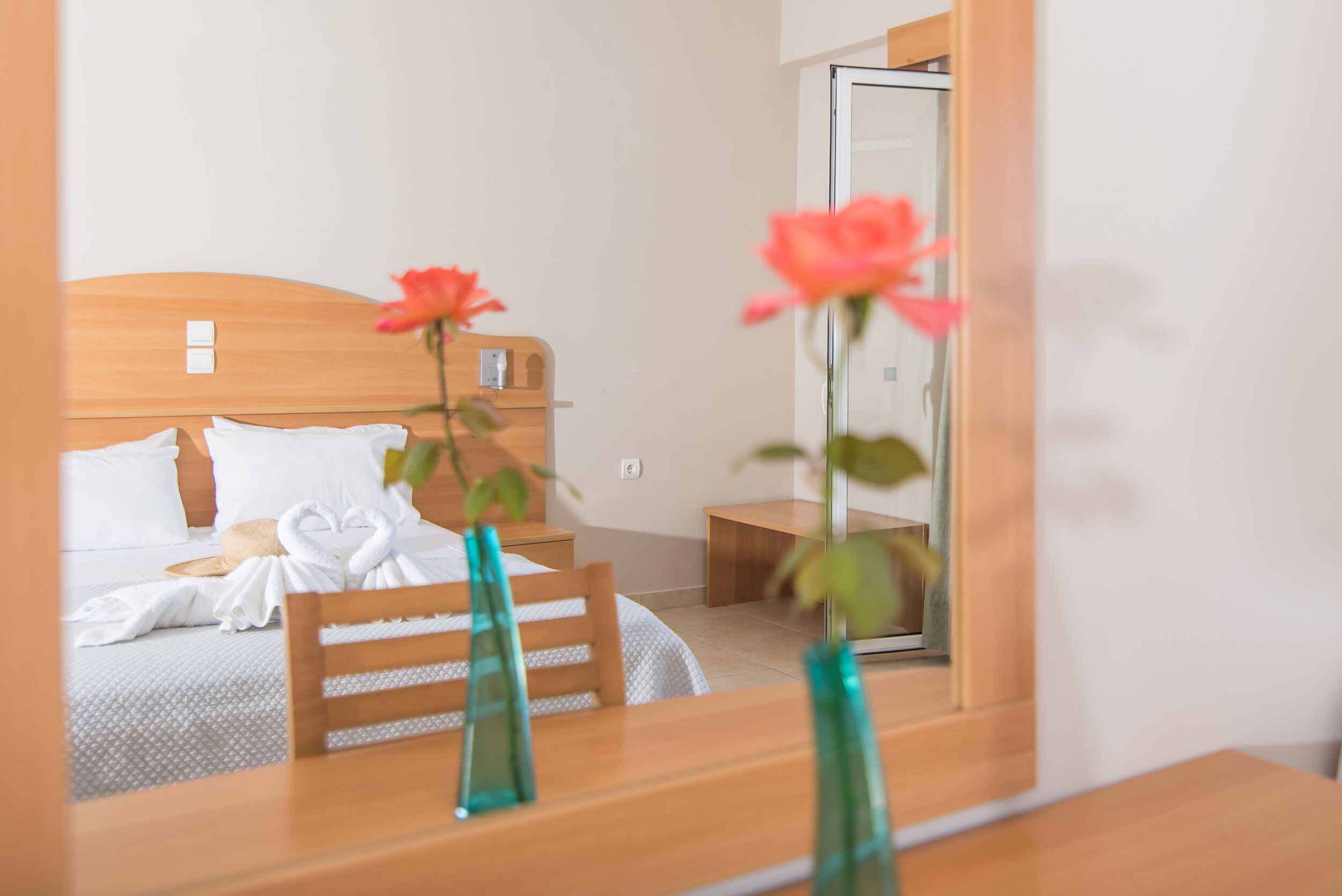 A hotel room with a wooden bed, white linens, a wooden nightstand, and a mirror reflecting a pink flower.