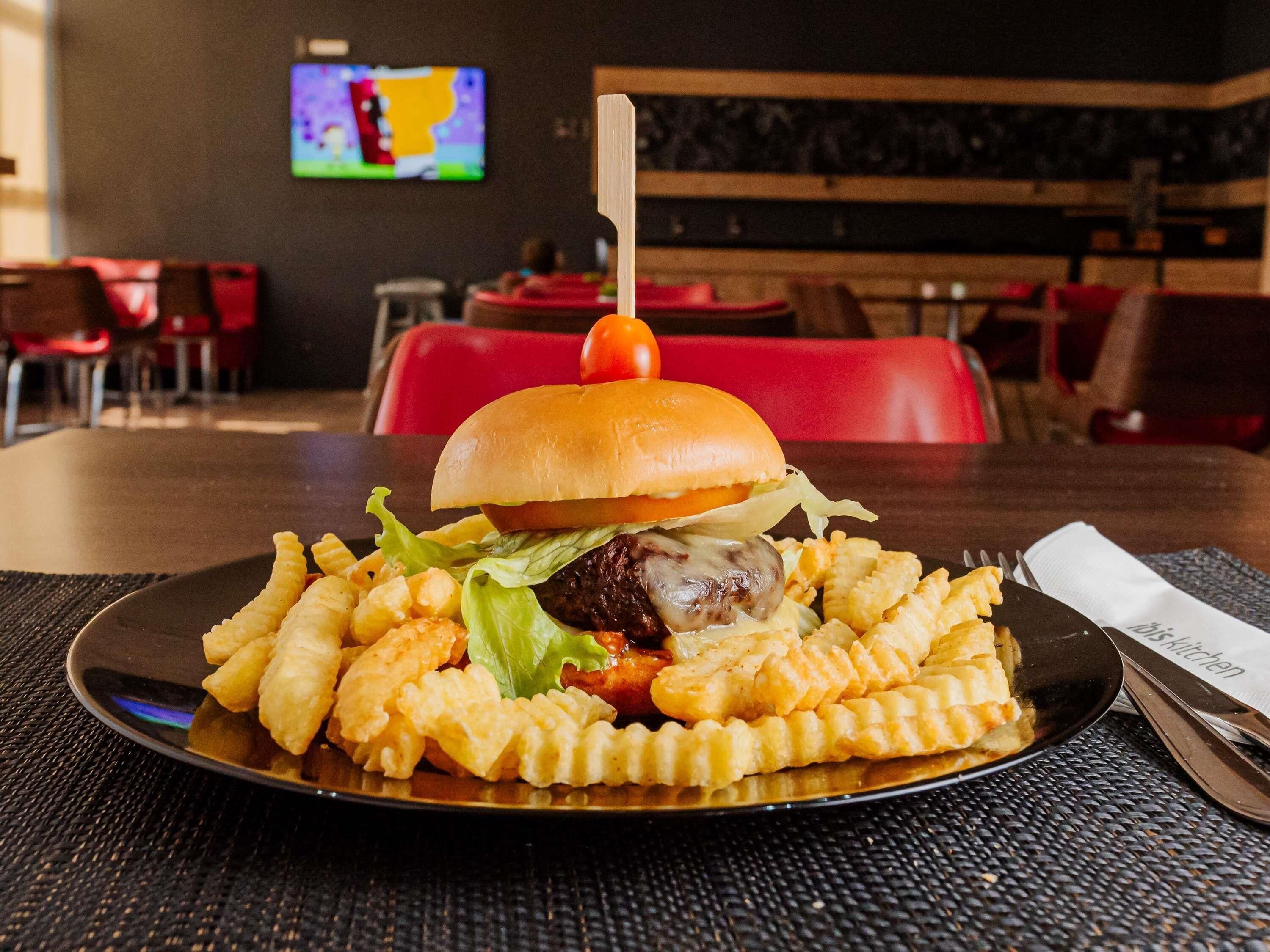 Un repas composé d’un hamburger maintenu par un cure-dent, de frites et d’une petite tomate, le tout présenté sur une assiette noire, posée sur une table recouverte d’un napperon tissé.