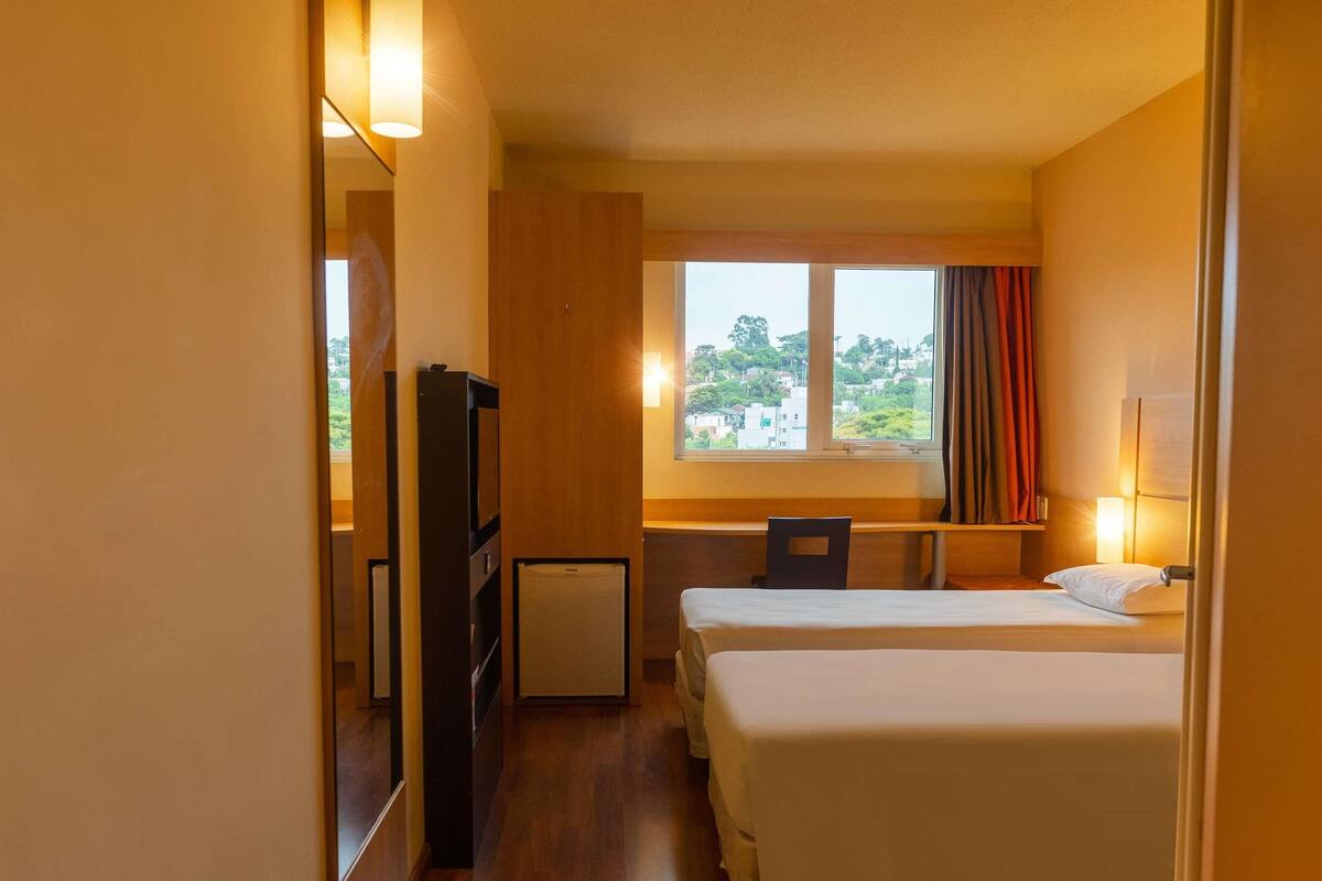 A hotel room with two beds, a desk, a TV, and a window with a view of buildings.