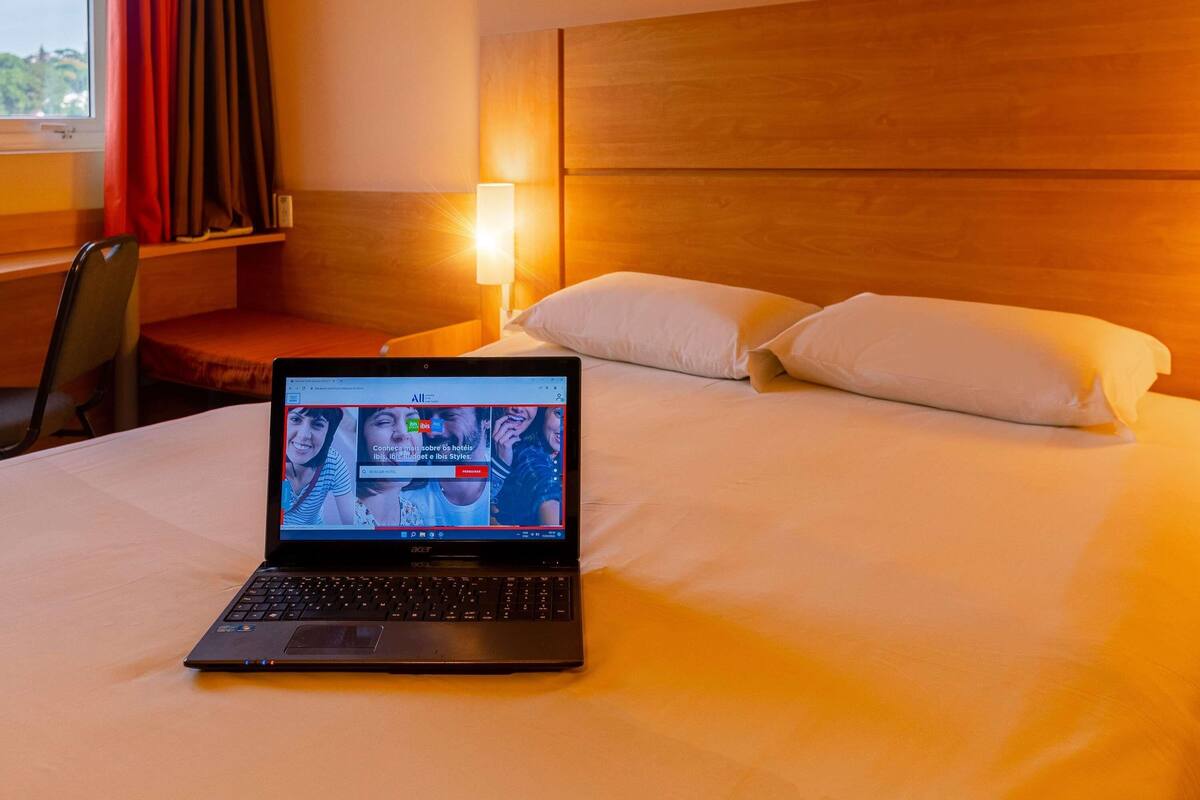 A laptop on a bed with a wooden headboard and a nightstand with a lamp.