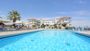 A swimming pool with lounge chairs and umbrellas, surrounded by buildings with red-tiled roofs. - Hilltop Hotel (Kassandra)