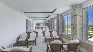 A corridor with wicker chairs and tables, large windows, and a stone wall. - Hilltop Hotel (Kassandra)