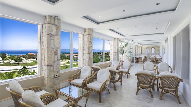 A spacious indoor area with wicker chairs and tables, large windows offering a view of the sea and distant landscape. - Hilltop Hotel (Kassandra)