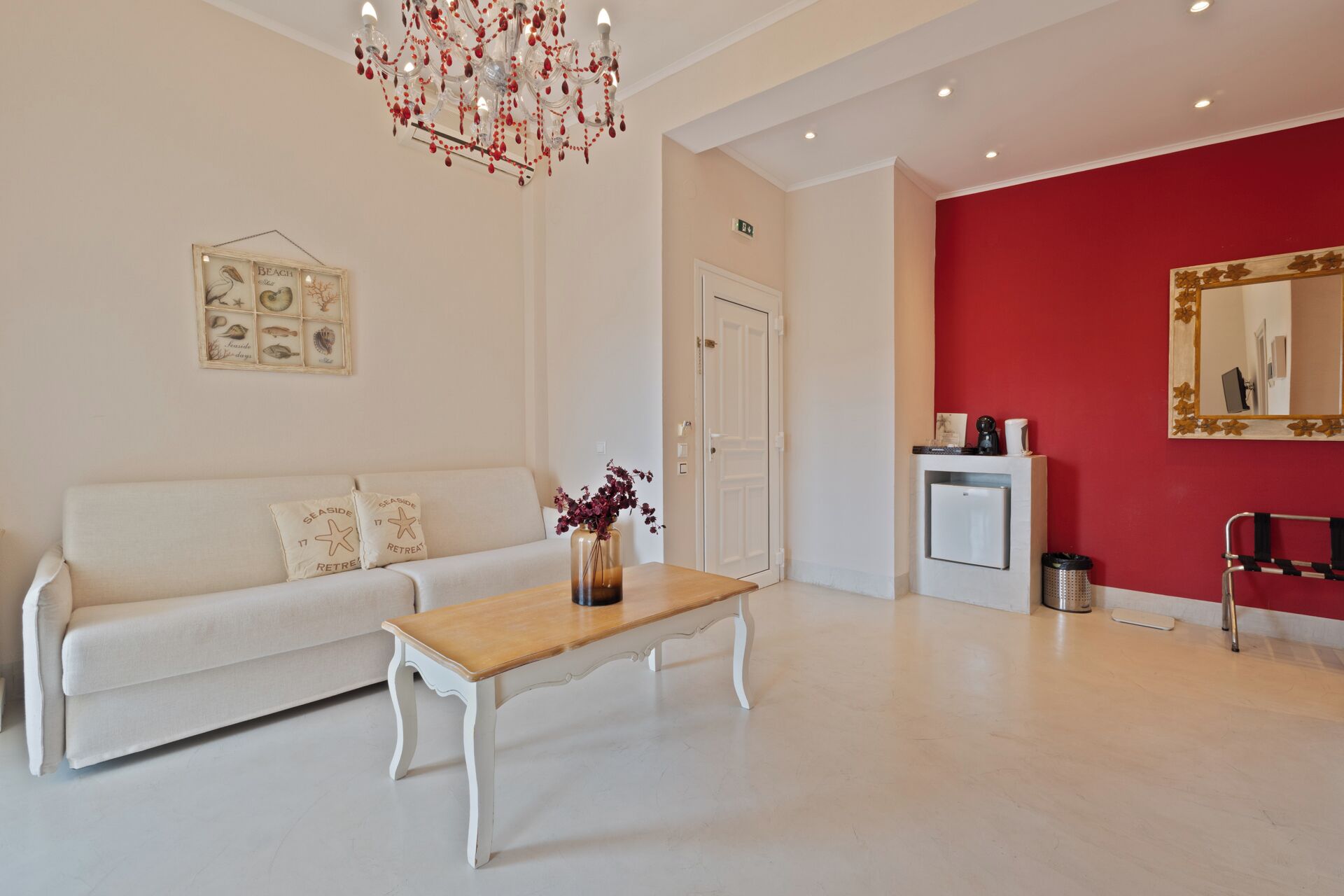 A modern hotel lobby with a white sofa, a wooden coffee table, a red accent wall, a small refrigerator, and a mirror.