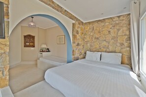 Un dormitorio con pared de piedra, una cama con ropa blanca, cabecera de madera y una araña.