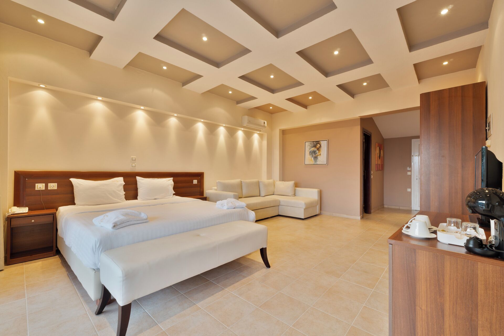 A spacious hotel room with a large bed, a sofa, a desk with a computer, and a ceiling with recessed lighting.