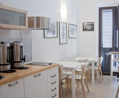 Una cucina moderna con mobili bianchi, elettrodomestici in acciaio inossidabile e un piano di lavoro in legno. Si notano tavoli e sedie da pranzo bianche, e quadri appesi alle pareti.