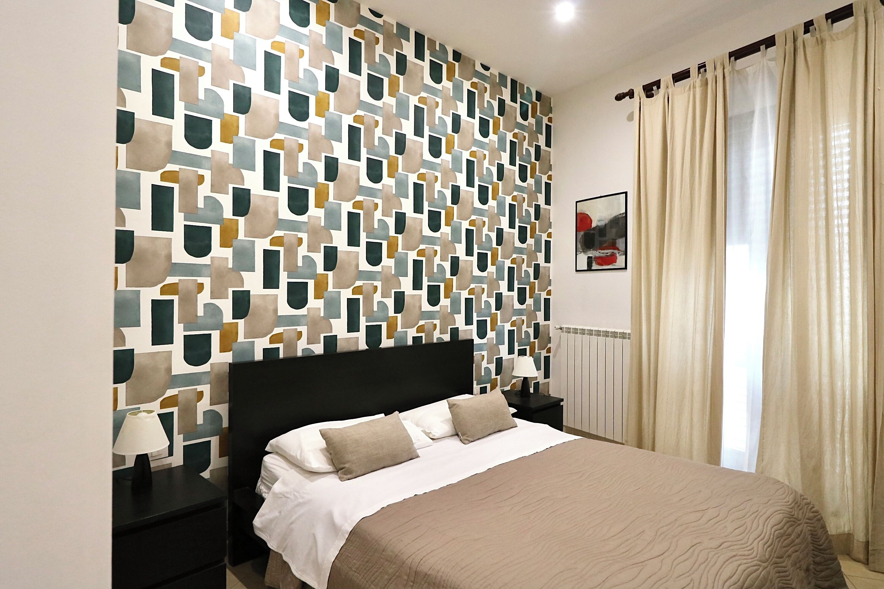 A neatly arranged bedroom with a patterned wall, a bed with a beige bedspread, and a nightstand with a lamp.