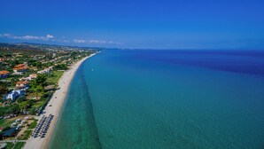 An aerial view of a coastal area with a long sandy beach, clear blue waters, and a mix of residential buildings along the shore. - Flegra Palace (Kassandra)