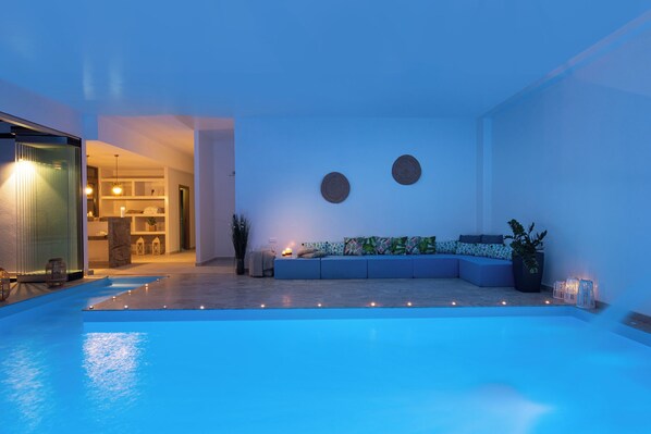 Un espace piscine intérieur moderne, avec une piscine illuminée de lumière bleue, un coin salon doté de coussins et un mur orné de deux motifs circulaires.
