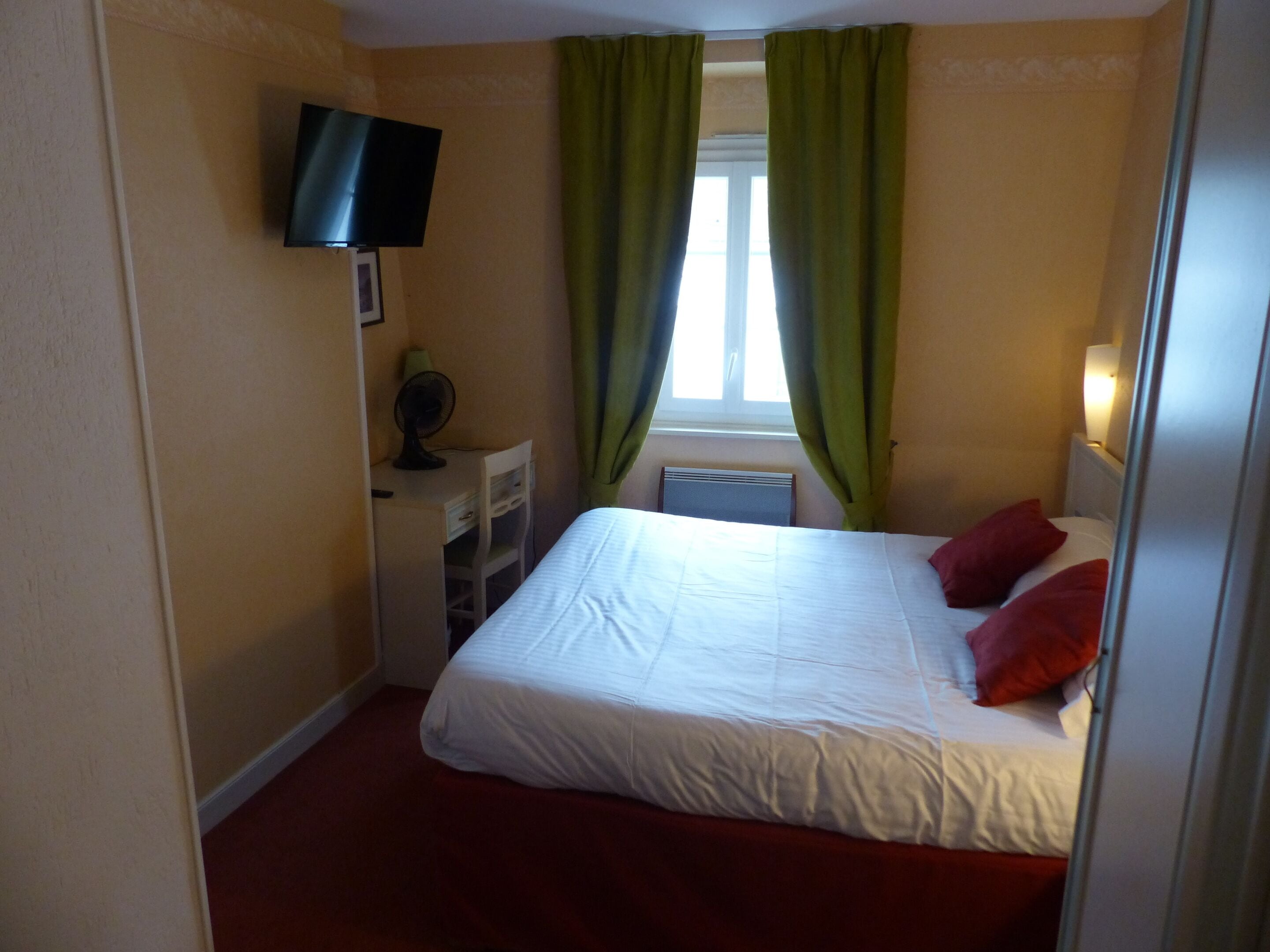 A bedroom with a bed, a desk, a chair, a television mounted on the wall, and a window with curtains.