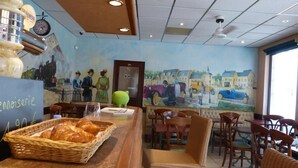 A restaurant interior with a mural, wicker chairs, a counter with pastries, and a clock. - Hôtel Belle Epoque (Chinon)
