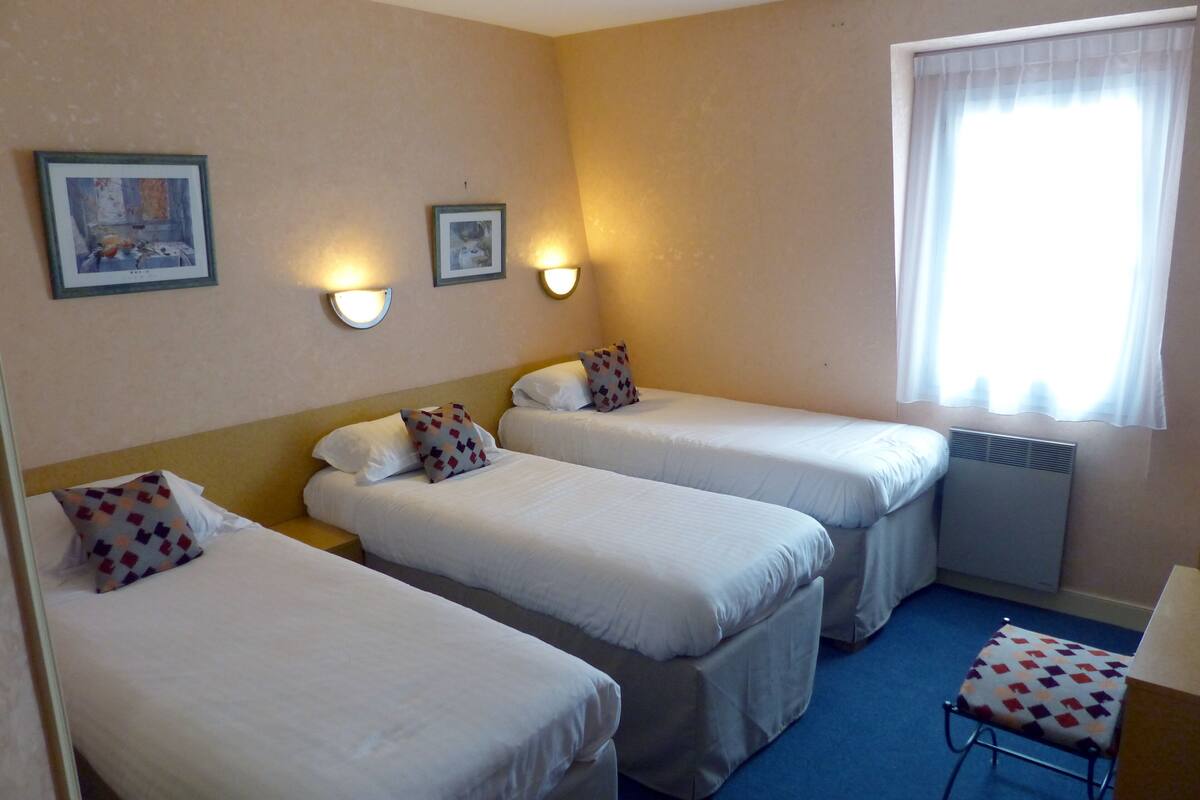 A hotel room with three single beds, a small table, and two wall-mounted lamps.