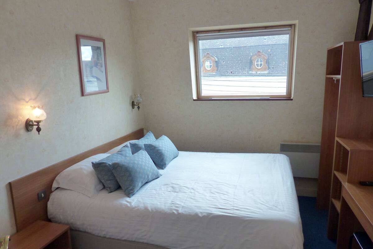 A neatly made bed with pillows, a bedside table, a wall-mounted lamp, a window with a view, and a wooden wardrobe.