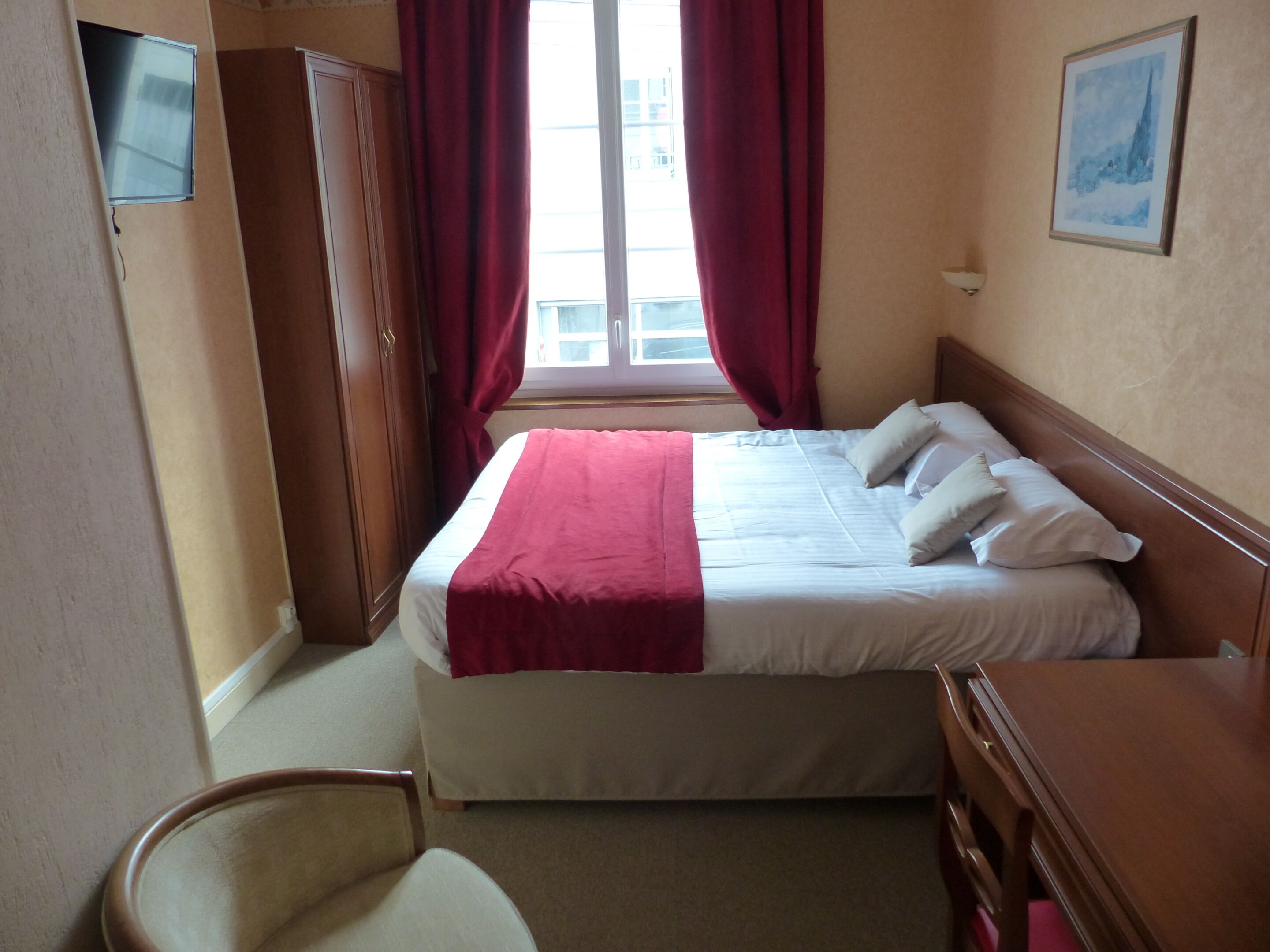 Habitación de hotel con cama, una silla, escritorio, ventana con cortinas y un cuadro en la pared.
