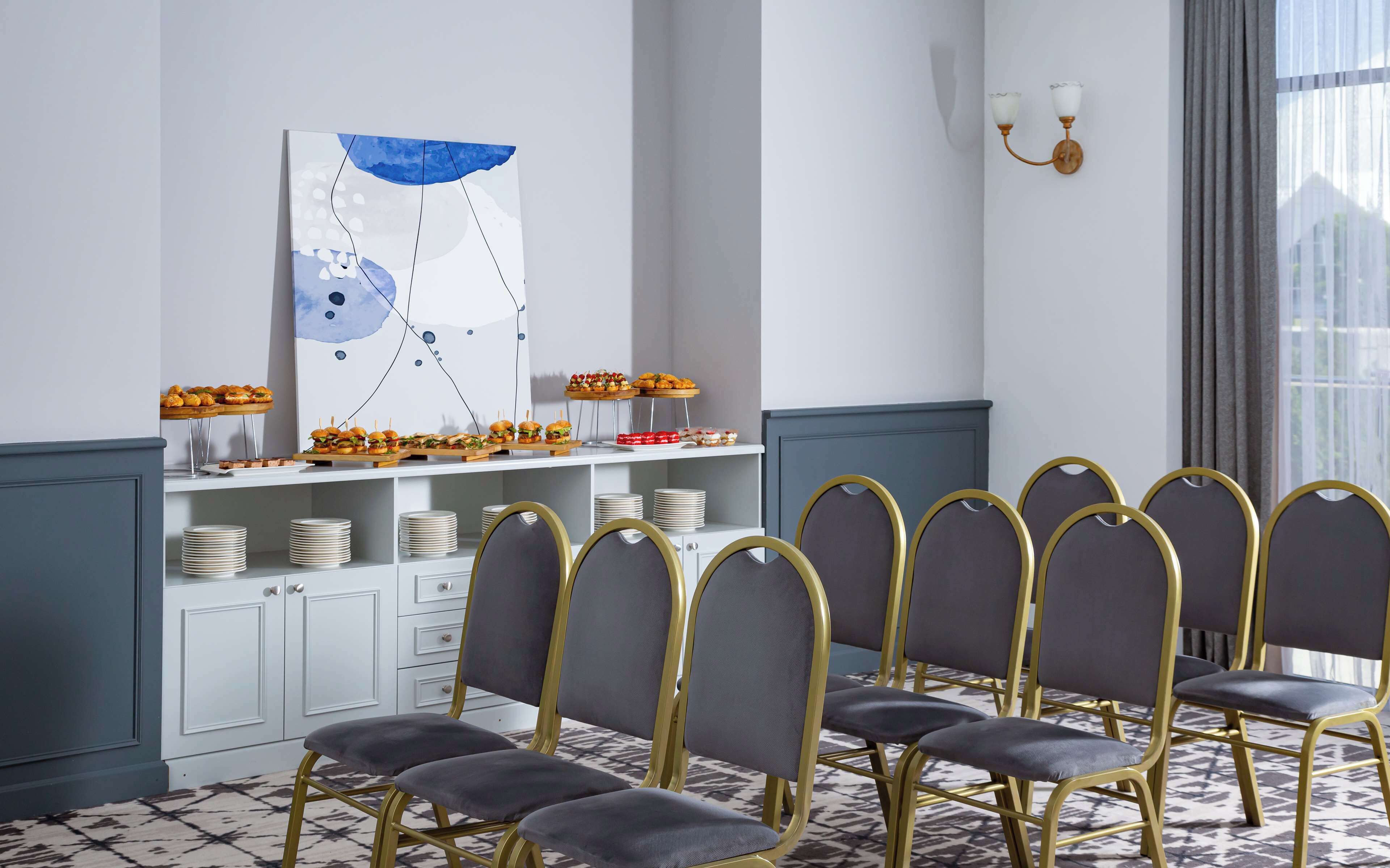 Un comedor con mesas de buffet, un cuadro abstracto grande y una fila de sillas con marcos dorados.