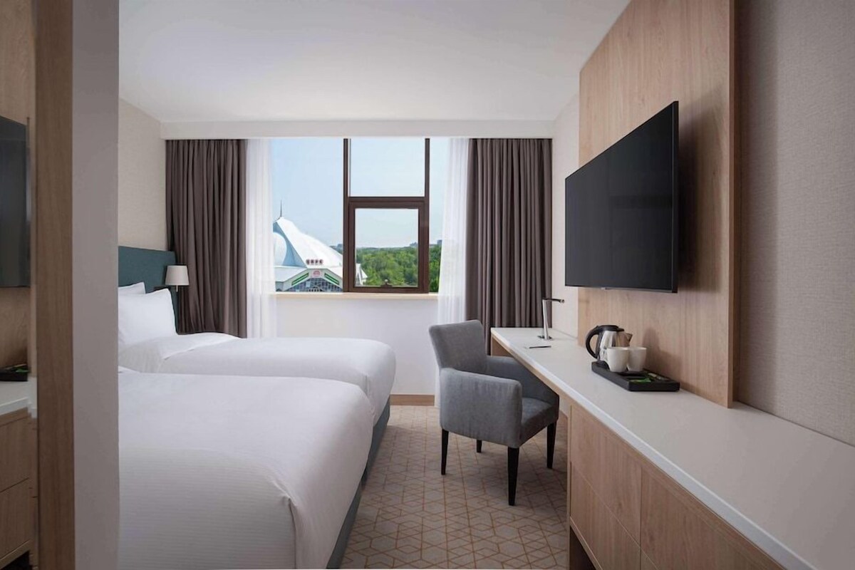 A hotel room with two beds, a desk, a chair, a television, and a window with a view of a building.