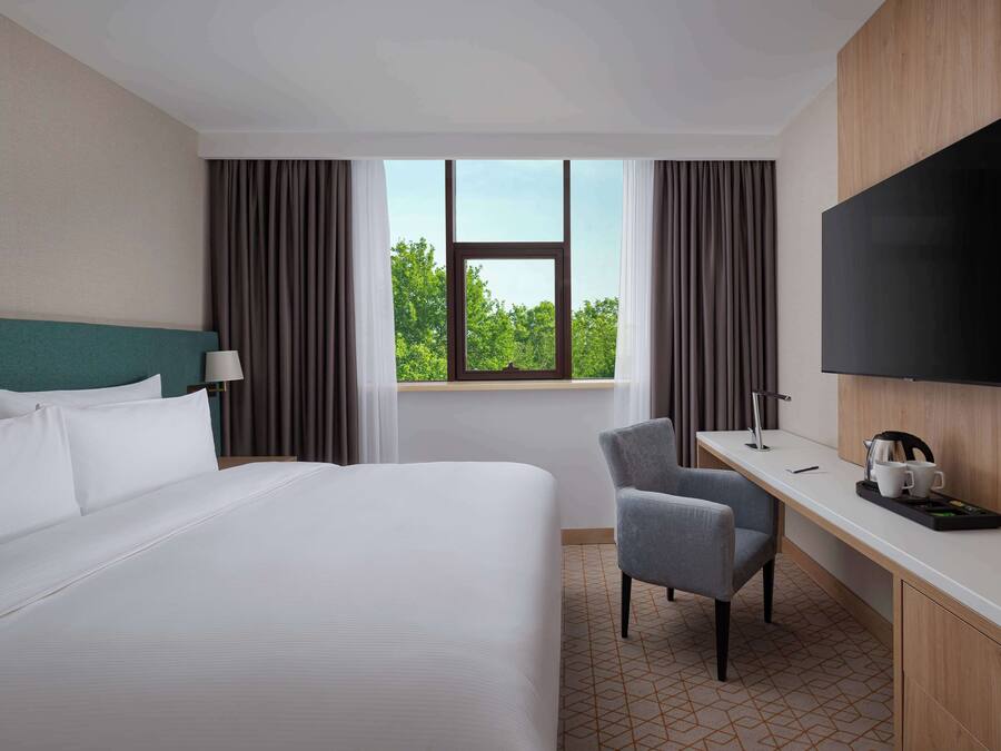 Ein Hotelzimmer mit einem großen Bett, einem Schreibtisch mit Stuhl, einem Flachbildfernseher und einem Fenster mit Vorhängen.