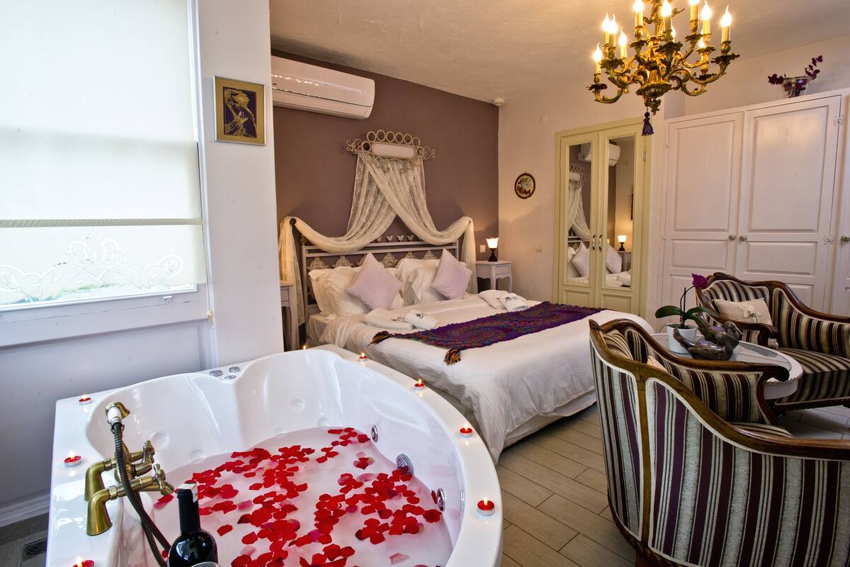 A bedroom with a large white bathtub filled with rose petals, a bed with white linens, a chandelier, and striped upholstered chairs.