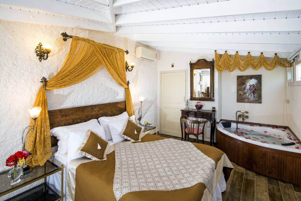A bedroom with a large bed, a wooden headboard, a bedside table with a lamp, a nightstand with a vase of flowers, a bathroom with a bathtub, a mirror, and a door.