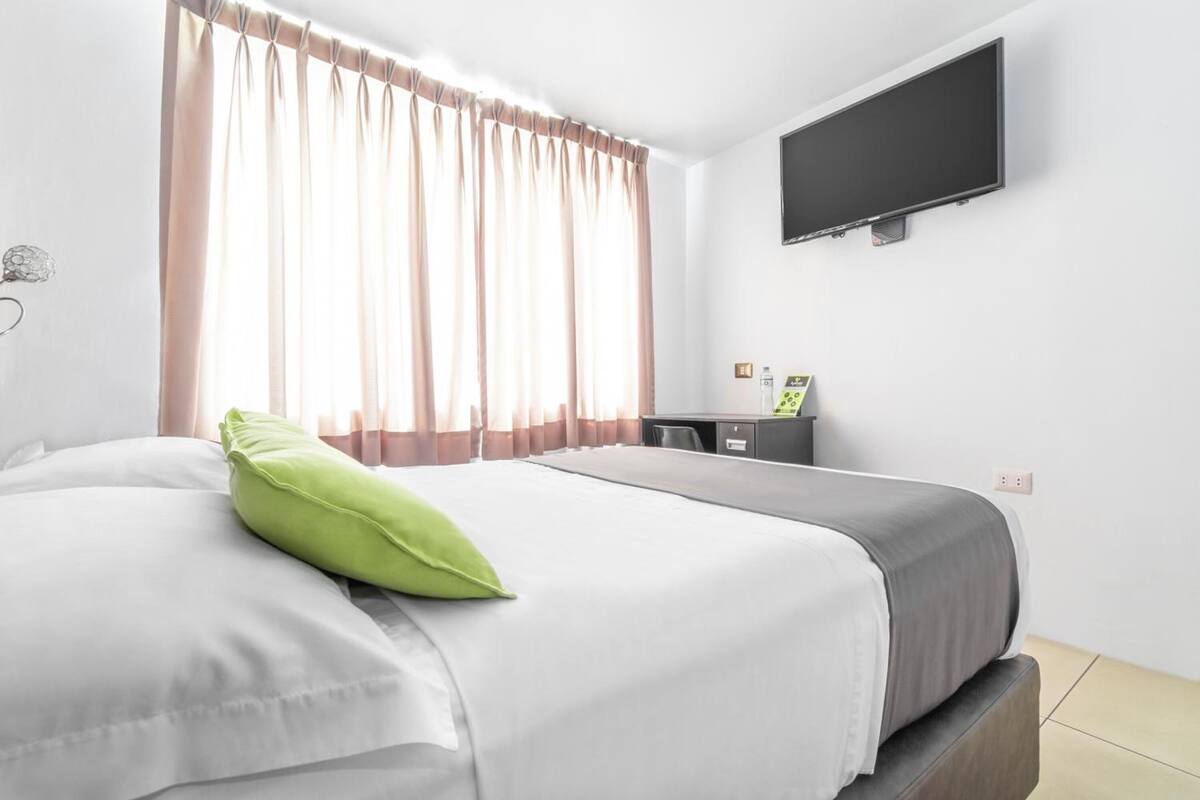 A hotel room with a bed, a desk, a flat-screen TV mounted on the wall, and a window with curtains.
