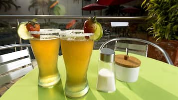 Dos vasos altos de una bebida amarilla, adornados con lima y fresas, un salero y un recipiente de madera sobre una mesa verde al aire libre.