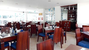 Un restaurante con varias mesas y sillas, algunas con cojines azules. Hay copas y botellas de vino sobre las mesas. El restaurante tiene un diseño moderno con muebles de madera y un separador de vidrio.