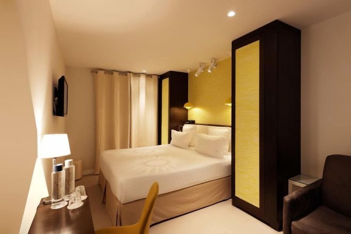 A hotel room with a bed, a chair, a desk with a lamp, and a wall-mounted TV.