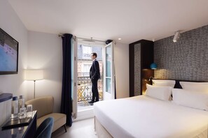 A hotel room with a bed, a desk with a chair, a table with glasses and a bottle, and a balcony with a person standing. - Hotel Eden (Paris)
