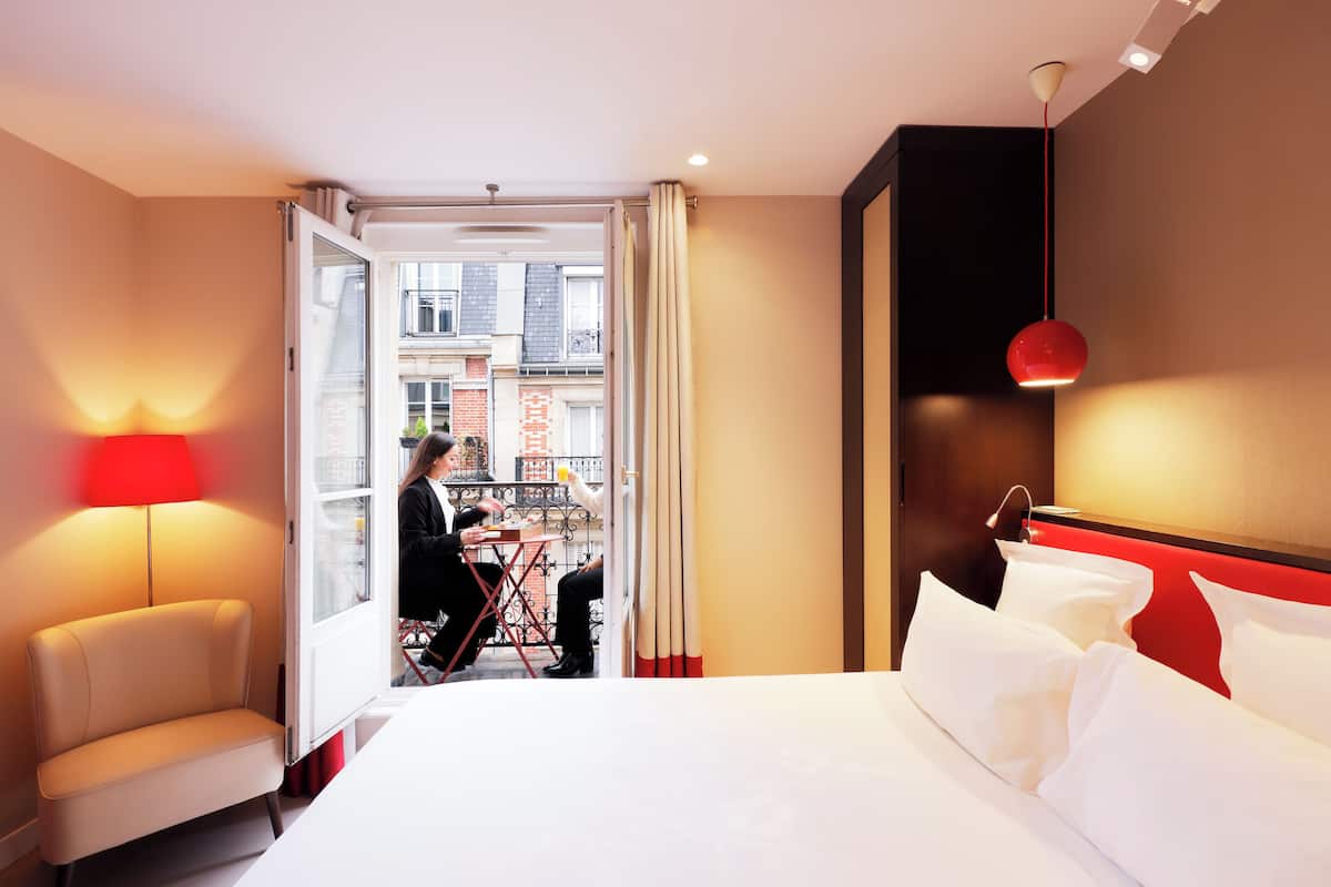 Habitación de hotel con cama, una silla, una mesa y balcón con vista a edificios.