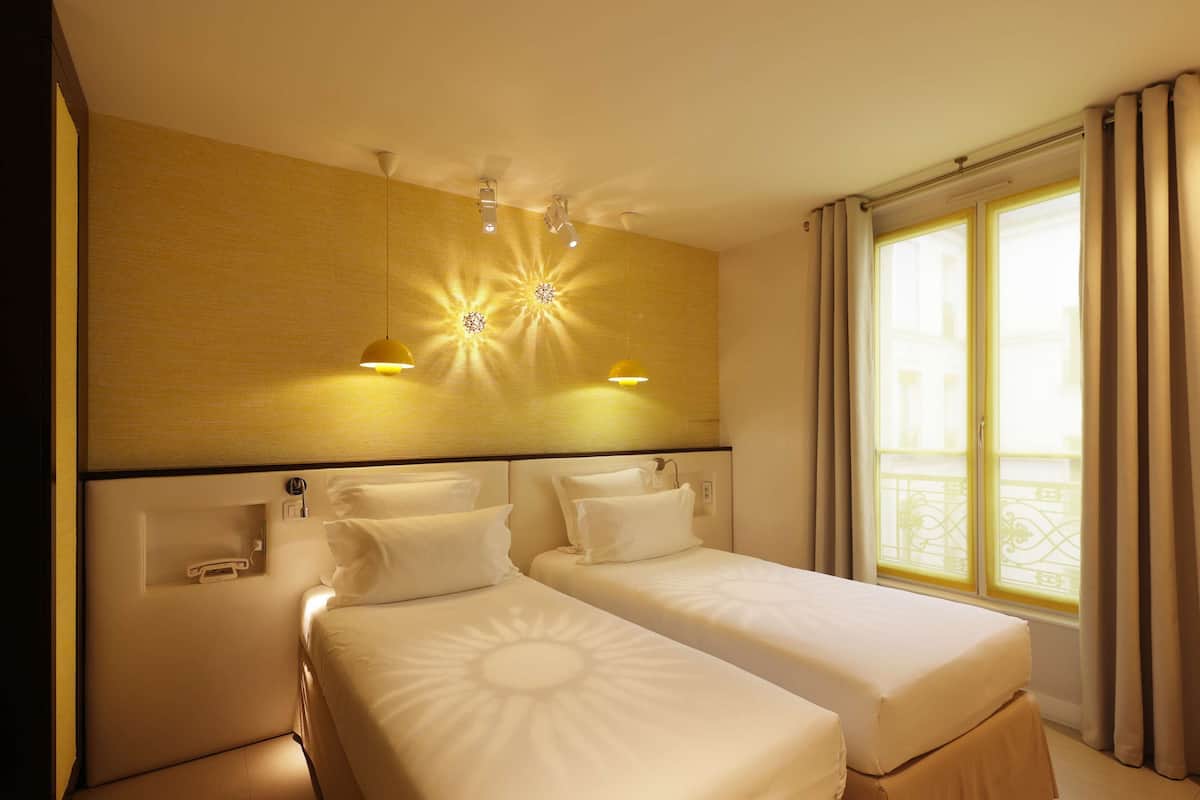 Habitación de hotel con dos camas individuales, una mesita de noche pequeña con un teléfono, una lámpara de pared y un ventanal con cortinas.