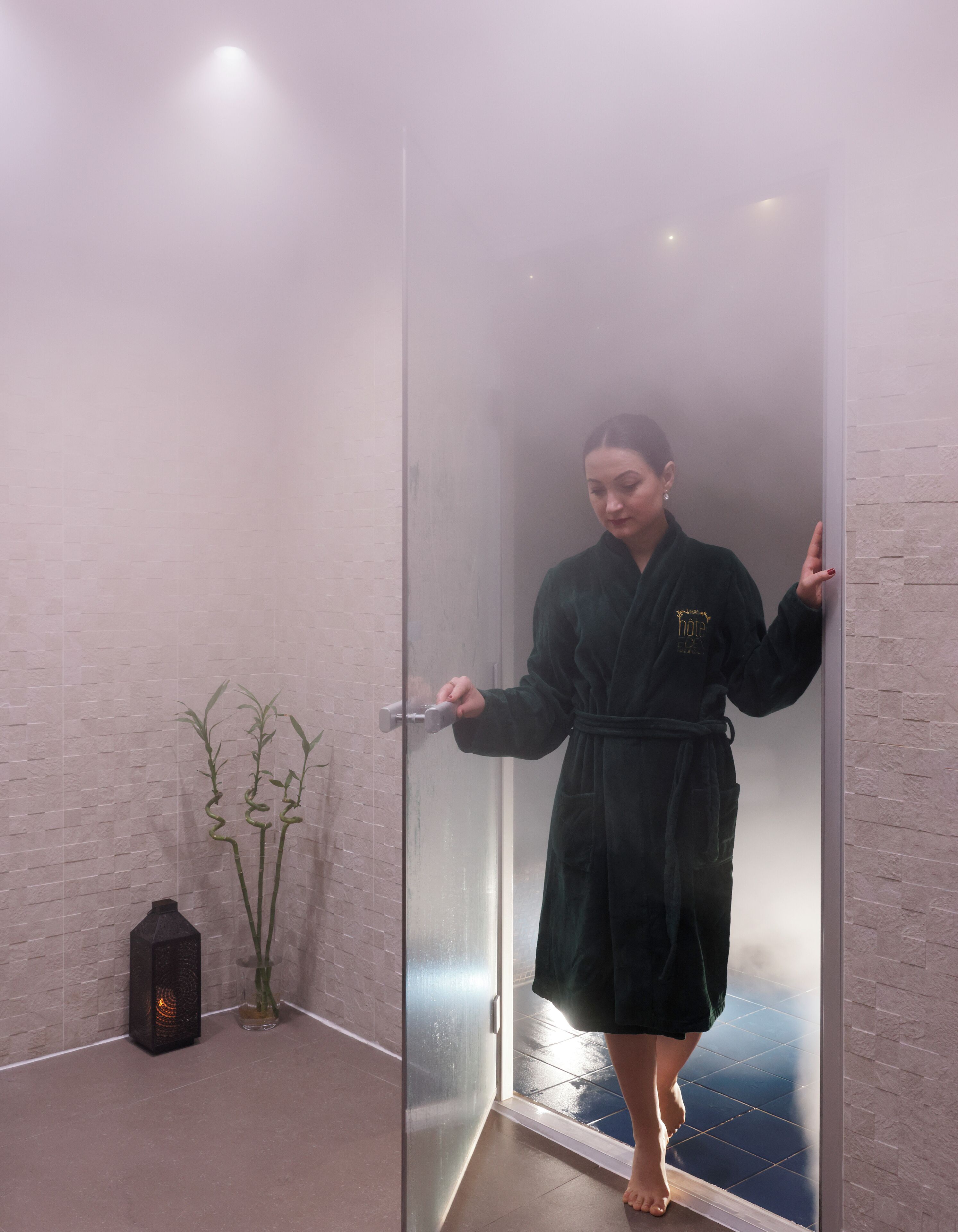 Una persona vestida con bata de baño en una habitación con puerta de vidrio, pared de ladrillo y una planta pequeña.