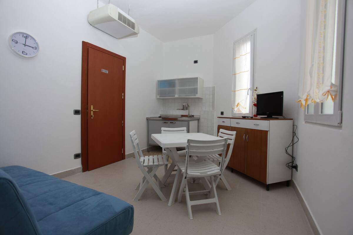 Ein kompakter Raum mit einem Bett, einem Esstisch mit Stühlen, einer kleinen Küchenzeile und einem Fernseher.