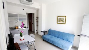 Uma sala de estar com um sofá azul, uma mesa de jantar com cadeiras, uma mesinha com uma planta e uma geladeira branca.