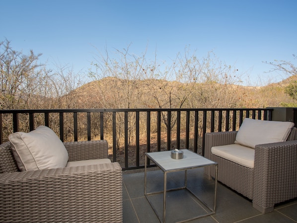 A balcony with wicker furniture, a metal table, and a view of a dry landscape. - Shepherd's Tree Game Lodge (Pilanesberg National Park)