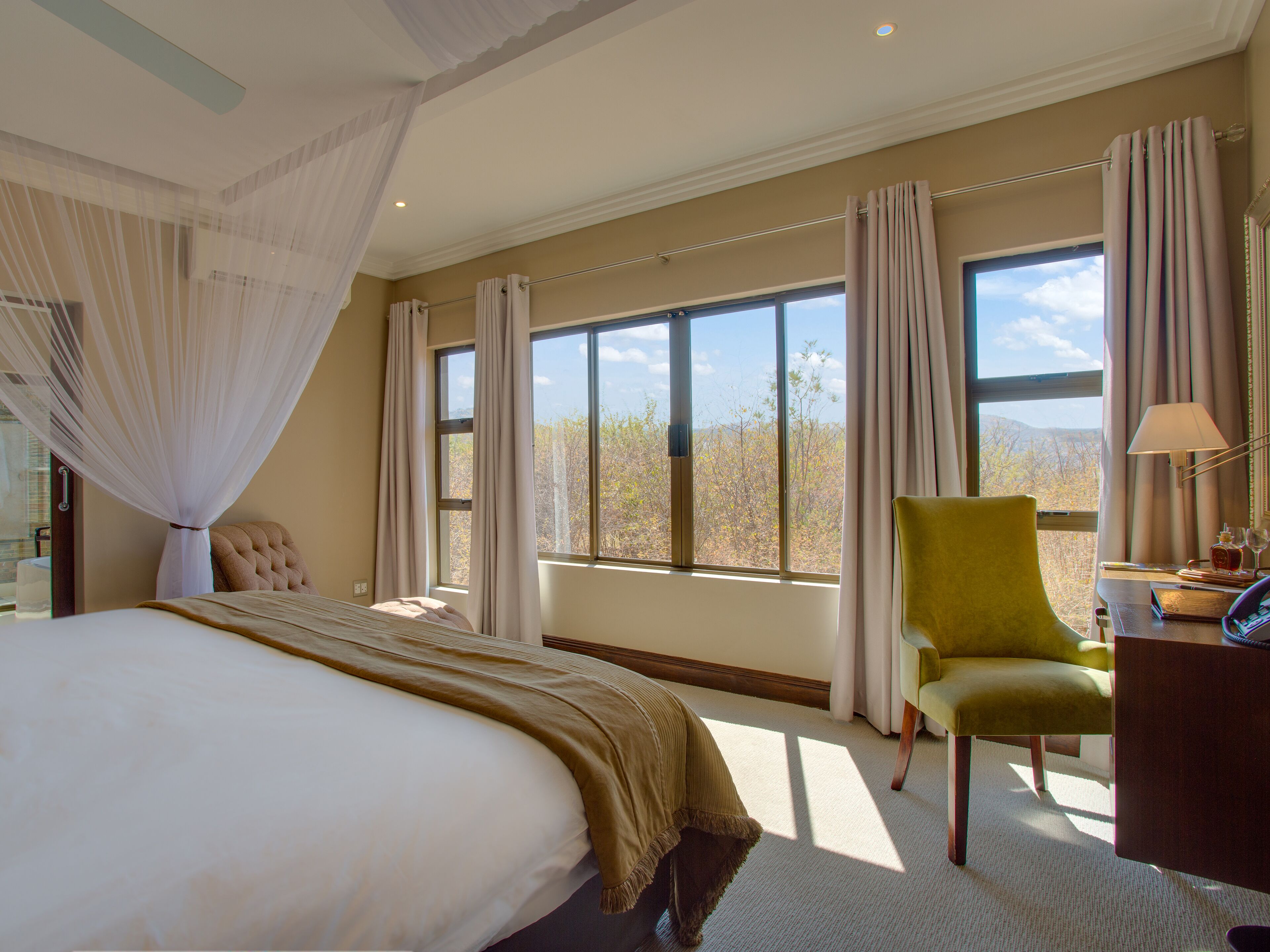 A bedroom with a large bed, a green armchair, a desk with a lamp, and a view of trees through the windows.