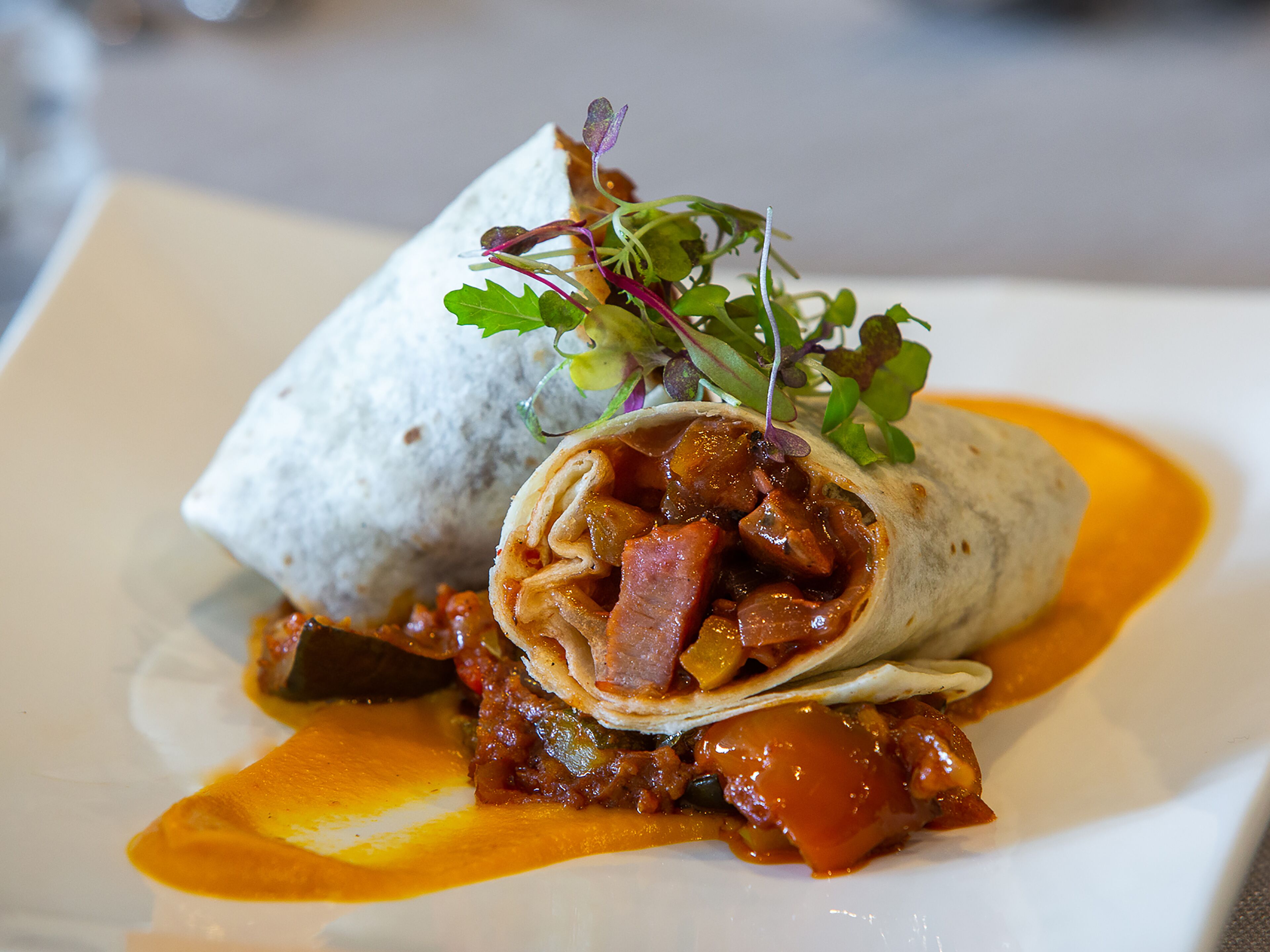 A burrito with a side of sauce and greens on a plate.