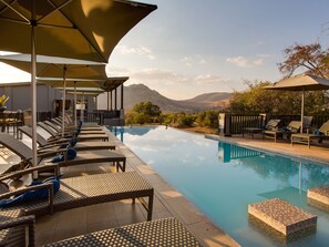 A poolside area with lounge chairs and umbrellas, a clear view of mountains in the background, and a calm pool. - Shepherd's Tree Game Lodge (Pilanesberg National Park)