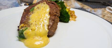 A steak with sauce on a plate, a glass of water, and a fork on a table.