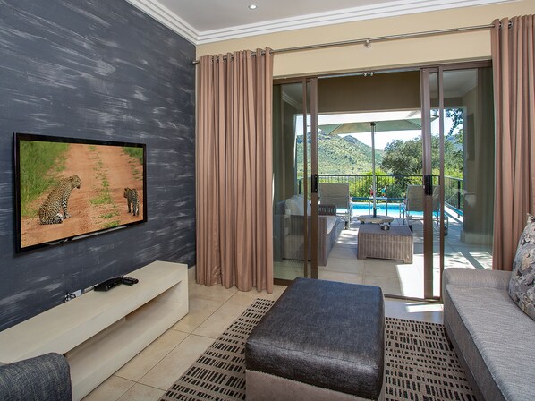 A modern living room with a flat-screen TV, a sofa, an ottoman, and a sliding glass door leading to a balcony with a pool. - Shepherd's Tree Game Lodge (Pilanesberg National Park)
