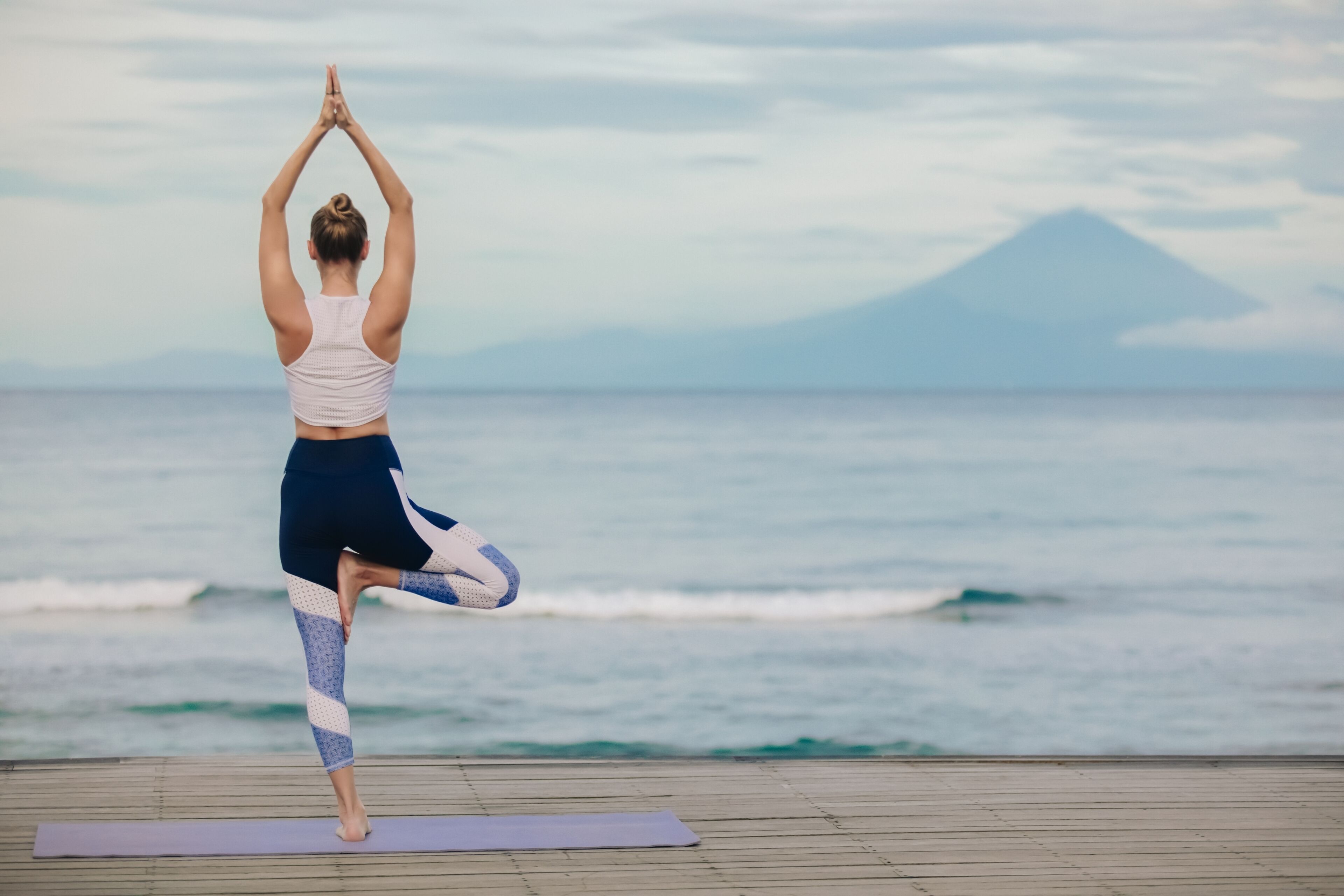Una persona realizando una postura de yoga sobre una esterilla frente al océano, con una montaña al fondo.