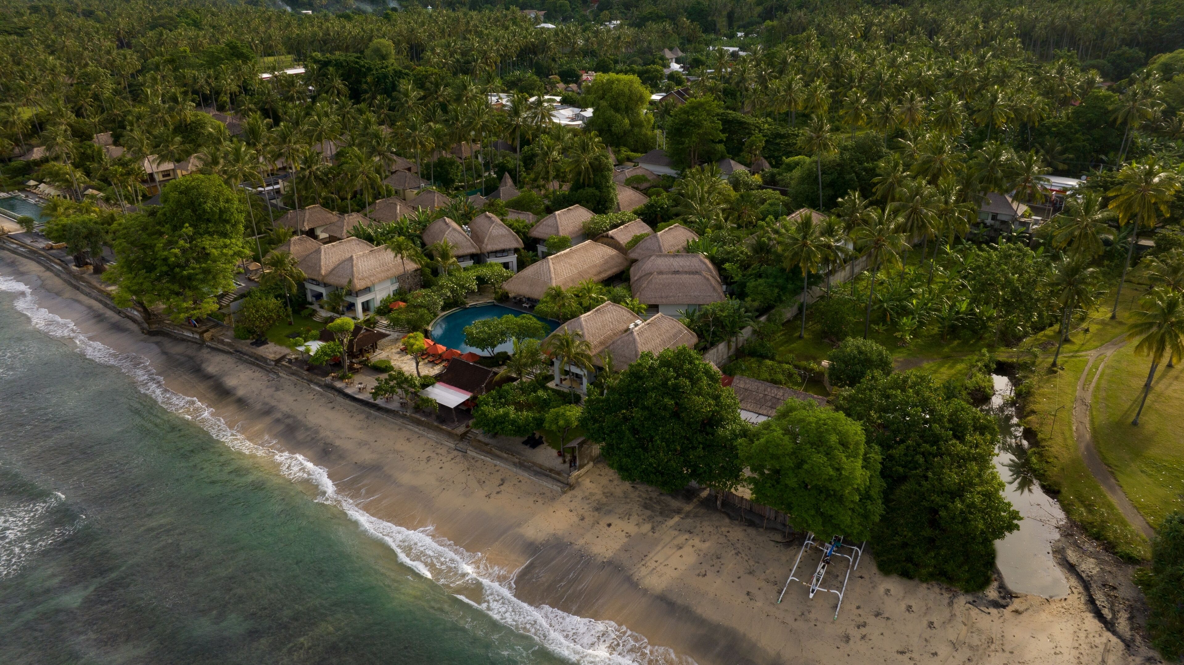 An aerial view of a resort with multiple thatched-roof bungalows, a swimming pool, and a beach.