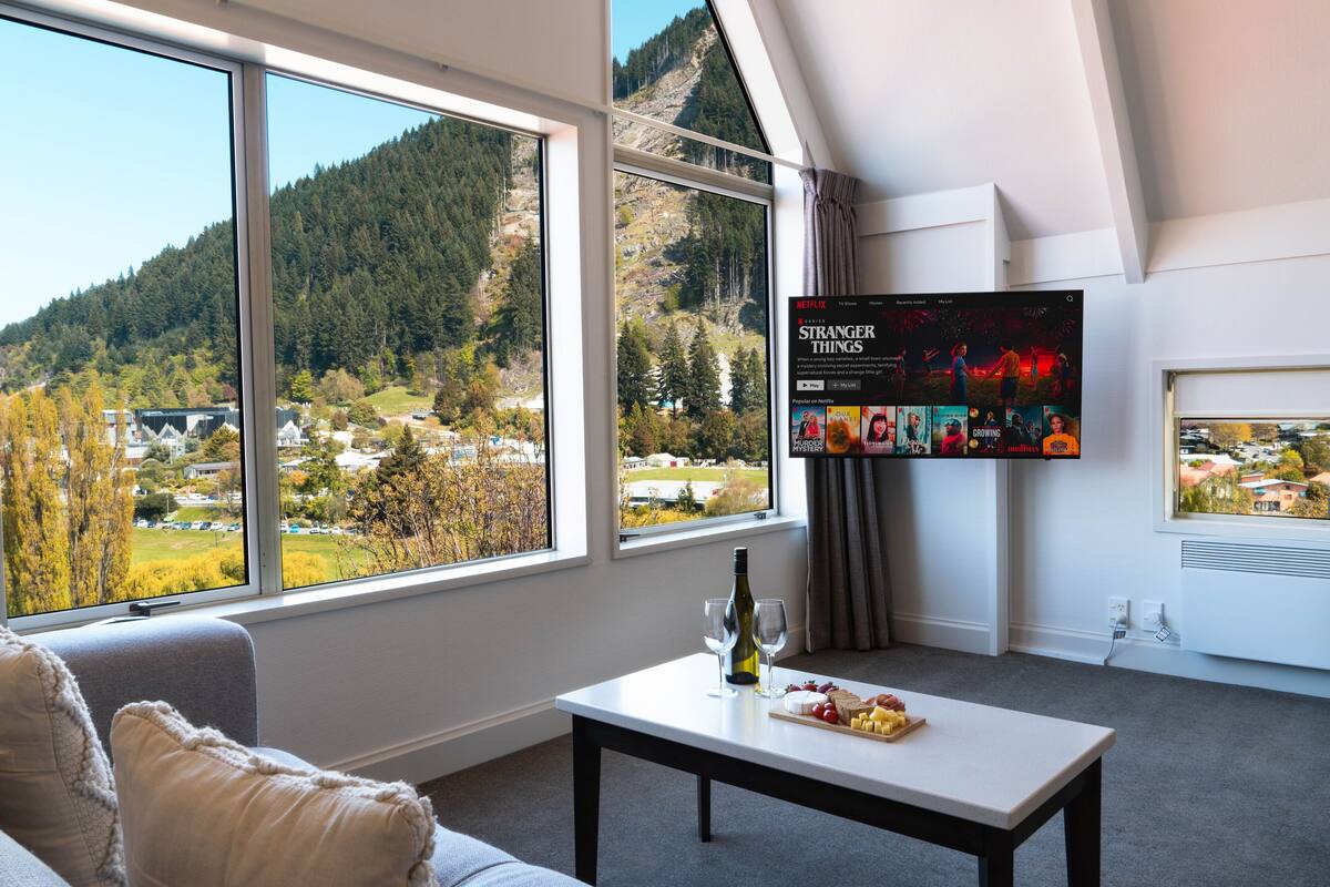 A modern living room with a large window offering a scenic view of a forested mountain and a town below. A flat-screen TV mounted on the wall displays a movie selection.